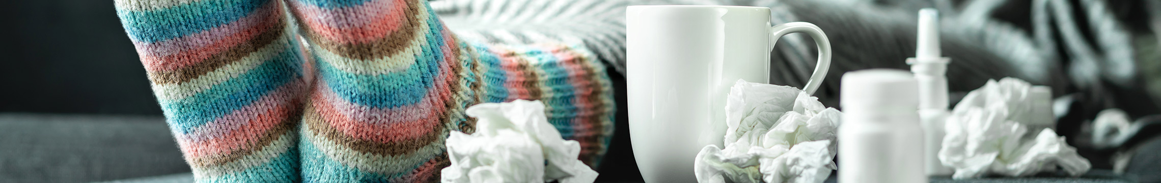 Woman on couch with cozy socks blowing nose while surrounded by tissues, medicine, and warm tea