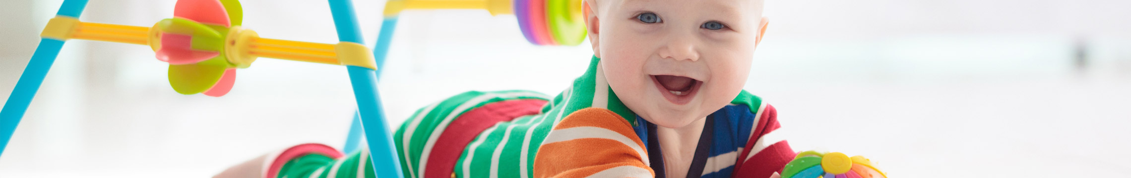 A smiling baby lays under a plastic play structure and plays with a colorful ball