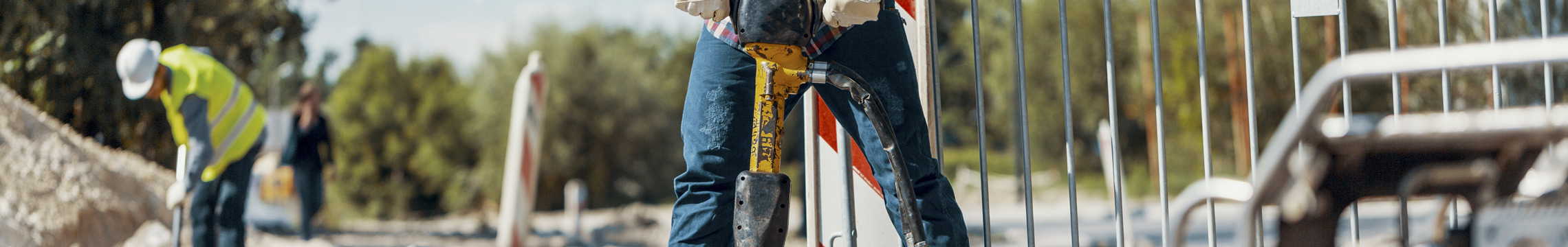 A road worker in jeans uses a jackhammer, in the distance another worker wearing a neon shirt is digging