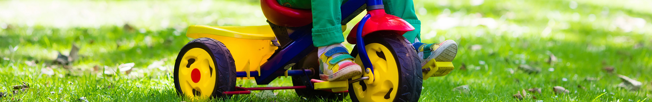 Little kid in multicolored outfit rides on a tricycle while eating a lollipop