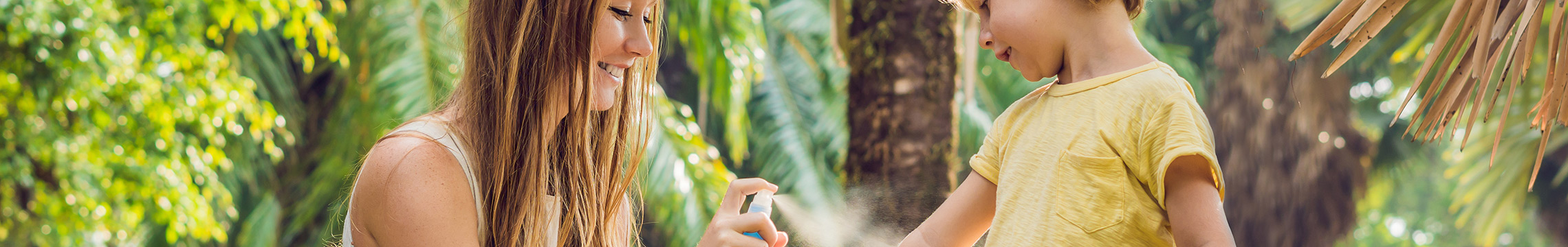 A smiling woman sprays bug spray on a young boy in a lush green outdoor setting