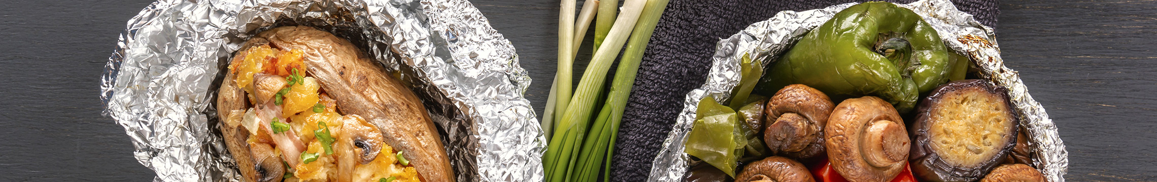 Baked potatoes and vegetables wrapped in foil on a dark table with utensils nearby